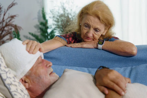 caucasian senior wife at sofa bed watching with concern to senior husband lying on bed at home with wet cloth cover forehead to reduce fever from sickness