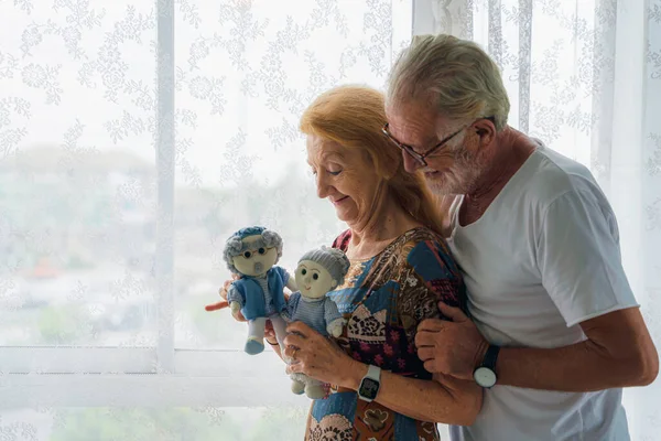 caucasian senior husband and wife couple standing by windowat home with old dolls on hand showing happy and romantic face of loving each other