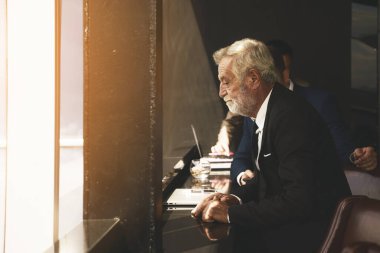 İş elbisesi içinde gri sakallı, beyaz saçlı beyaz iş adamının portresi iş yerinin penceresinin yanında duruyor.