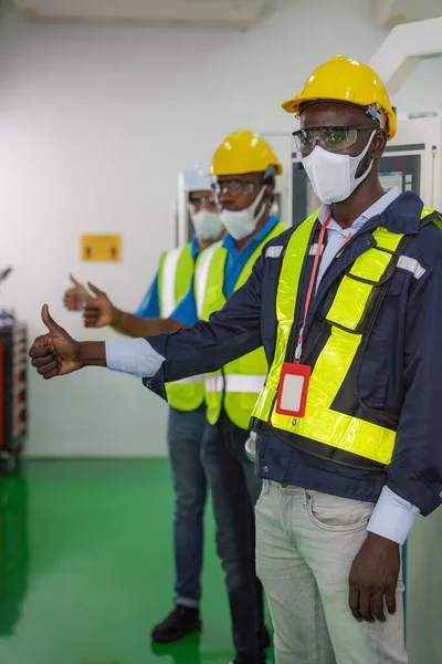 african american factory worker and team in uniform with safety vest and helmet wearing face mask for coronavirus protection thumb up together for teamwork in factory control room