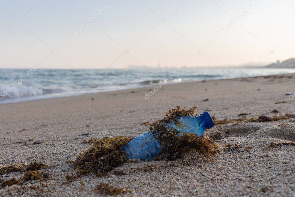 Blue plastic bottle covered in algae on the beach, with a mediterranean sea landscape background