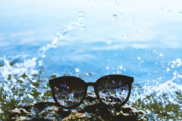 Occhiali da sole su una pietra su uno sfondo di un mare con spruzzi di — Foto Stock