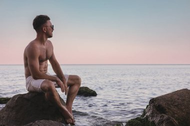 Bir adam kayanın üzerinde oturmuş denizin tadını çıkarıyor.