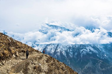 Nepal Himalayalar bir kayalık dağ yol boyunca iki turist yürümek. Karla kaplı dağlar bulutlar. 