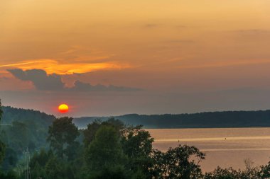 Bulutlu gökyüzü ve Volga Nehri kıyısında yeşil ağaçlar arka planı batan güneşin parlak sarı diski. 