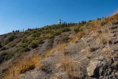 İnce selviler ve deniz feneri ile dik bir kayalık yamacı, Cri