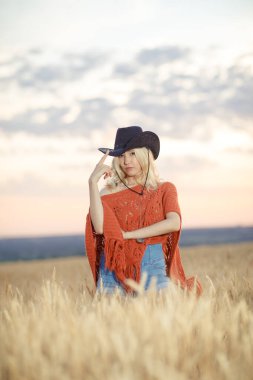 Genç bir kadın buğday tarlasında panço ve siyah şapka giymişti.