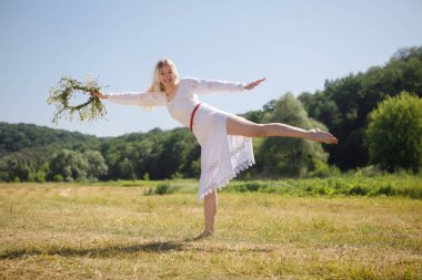 Beyaz elbiseli, uzun sarı saçlı genç beyaz kadın çelenkle tarlada dans ediyor.