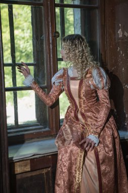 Orta çağ elbisesi giymiş beyaz kıvırcık saçlı genç kadın pencerenin yanında oturuyor.