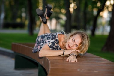 Sarı kıvırcık saçlı, elbiseli ve gözlüklü güzel bir genç kadın parkta yatıyor.