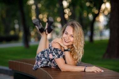 Sarı kıvırcık saçlı, elbiseli ve gözlüklü güzel bir genç kadın parkta yatıyor.