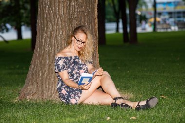 Sarışın, kıvırcık saçlı, elbise giyen ve gözlüklü güzel bir kadın parkta oturmuş kitap okuyor.