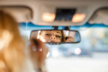 Sarışın, kıvırcık saçlı, takım elbiseli, gözlüklü, dışarıda arabada tabancalı ve aynada gülümseyen güzel bir genç kadın.