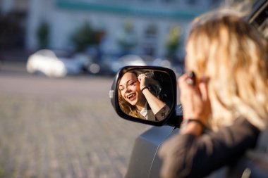 Sarışın, kıvırcık saçlı, takım elbiseli, gözlüklü, dışarıda arabada tabancalı ve aynada gülümseyen güzel bir genç kadın.