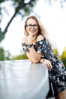 Sarışın, kıvırcık saçlı, güzel bir kadın. Parkta gözlüklü ve elbiseli.