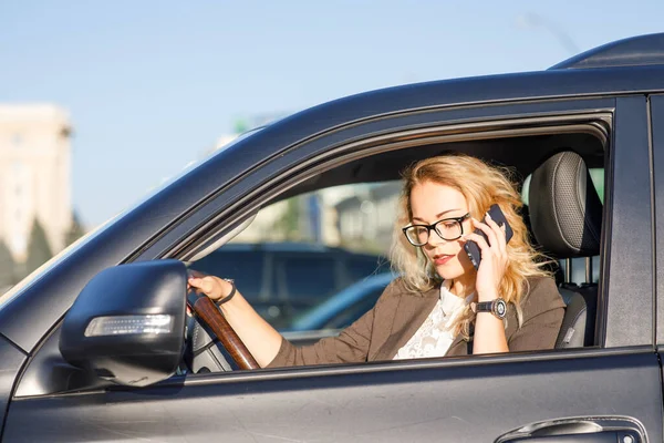 Beautiful young business woman with blond curly hair wearing a suit and glasses in a car talking by mobile phone