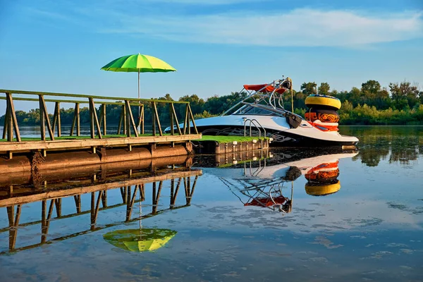 A small yacht is moored to the berth and ready to drive on the river. Tied inflatable rings are waiting for fun. Beach vacation. A green umbrella attached to the handrails protects from the sun. Mirror reflection in water.