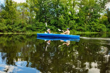 Yeşil ormanlarla çevrili sakin bir nehirde mavi bir teknede kano süren çift. Yaz macerası, aktif yaşam tarzı ve doğada su eğlencesi..