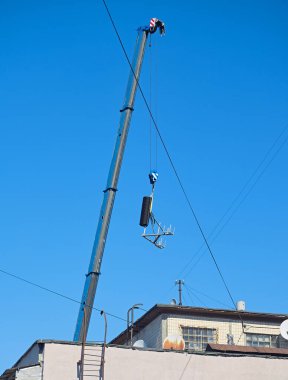 Açık mavi gökyüzüne karşı bir binanın çatısında vinç kaldırma ekipmanı, kablolar ve uydu antenleriyle endüstriyel çalışmalar.