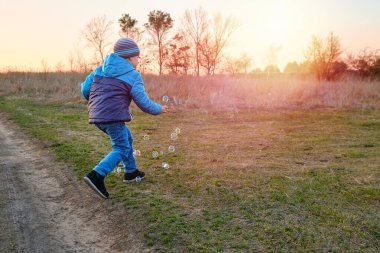 Çocuk kırsal bir yolda gün batımında dışarıda sabun köpüğü üflüyor. Kalın elbiseli çocuk altın saat boyunca oyun ve doğanın tadını çıkarıyor..