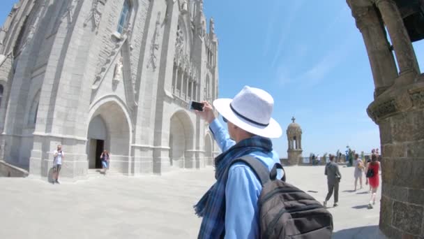2018 スカーフと帽子で若い男は スペインのバルセロナでティビダボの聖心の寺院の近くスマート フォンでビデオを撮影します — ストック動画