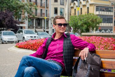 Genç gülümseyen adam, bir Bank Plaza Girona, İspanya turizm bir sırt çantası ile oturur