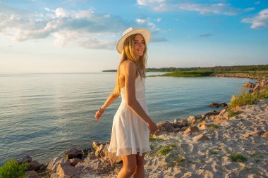 Beyaz yazlık saten elbise ve bir şapka giymiş güzel mutlu bir genç kadın deniz arka plan günbatımında fırtınası..