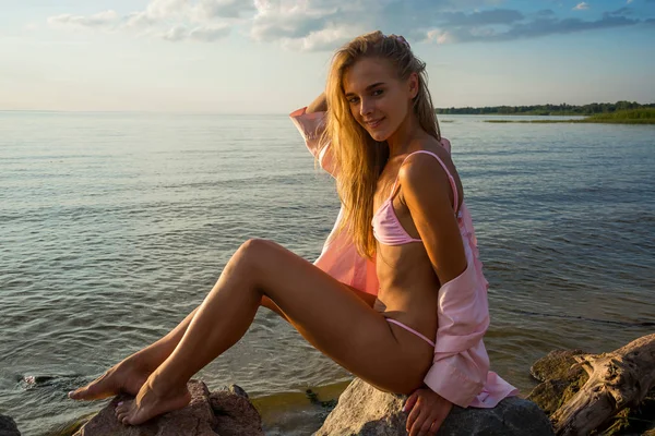 ピンクの水着を着た美しい幸せな若い女性とシャツは夕日の海の背景に座っています — ストック写真