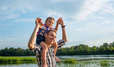 Mutlu genç baba onun oğlu Atçılık yukarıya bakıp gülerek onun omuzlarında tutar. Küçük oğlan sırtıma babası nehir arka plan, omuzlarında oturuyor