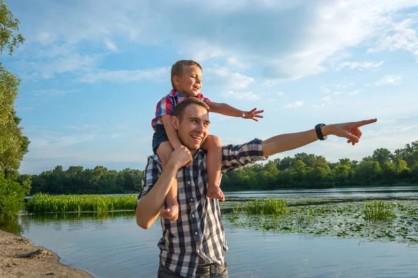 Happy young father holds his son piggyback ride on his shoulders, points at something with his hand and laughing. Little boy is sitting piggyback on shoulders his dad at river background