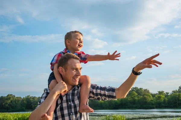 Happy young father holds his son piggyback ride on his shoulders, points at something with his hand and laughing. Little boy is sitting piggyback on shoulders his dad at river background