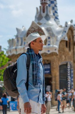 Genç bir adam bir beyaz standları şapka ve atkı sırt çantası ile yakın Guell Park, bir gingerbread ev içinde mimar Gaudi Barcelona, İspanya