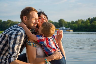 Onun baba ve anne küçük çocuk hugs ve onlar havada gülümseyen günbatımında bir nehir yakınında. Mutlu bir aile