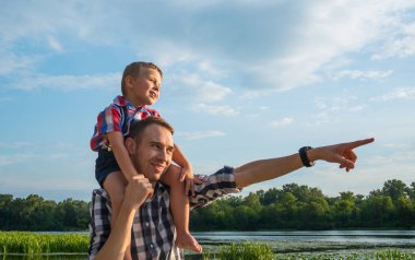 Mutlu genç baba onun oğlu Atçılık onun omuzlarında tutar, bir şey de eliyle işaret ve gülen. Küçük oğlan sırtıma babası nehir arka plan, omuzlarında oturuyor