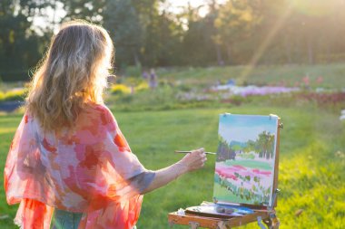Bir genç kadın sanatçı bir geri tutan bir fırça ve boya bir resim güneşin ışınları bir şövale üzerinde. Ressam yağlıboya tablolar bahçede günbatımında boyar.