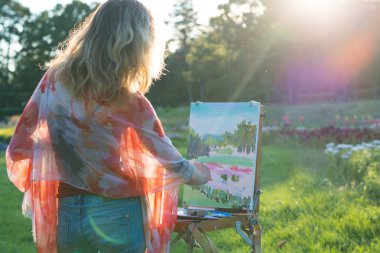 Bir genç kadın sanatçı bir fırça tutar ve güneşin ışınları bir şövale resmi boyar. Ressam yağlıboya tablolar bahçede günbatımında boyar.