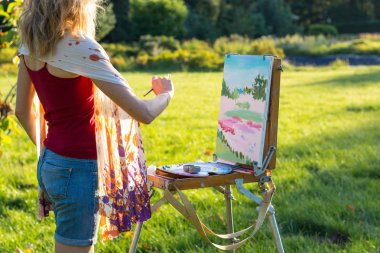 Bir genç kadın sanatçı bir fırça tutar ve güneşin ışınları bir şövale resmi boyar. Ressam yağlıboya tablolar bahçede günbatımında boyar.