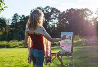 Bir genç kadın sanatçı bir fırça tutar ve güneşin ışınları bir şövale resmi boyar. Ressam yağlıboya tablolar bahçede günbatımında boyar.