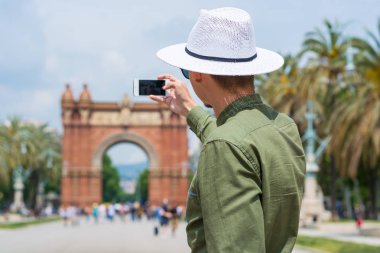 Genç bir adam, turist, şapkalı Catalonia, İspanya Barselona şehir Arc de Triomf yakınındaki bir akıllı telefon ile fotoğraf çeker