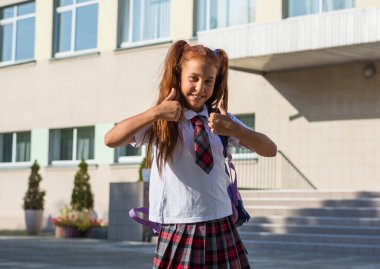 Gülümseyen ve okul bahçesi bir başparmak gösterilmesini bir sırt çantası ile Tekdüzen bir lisede bir kızla. Okula dönüş.
