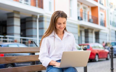 Genç güzel gülümseyen kadın bir dizüstü bilgisayar ile şehrin iş bölümünde bir bankta oturur. Genç güzel kadın, freelancer, yaz aylarında dizüstü bilgisayarda çalışır