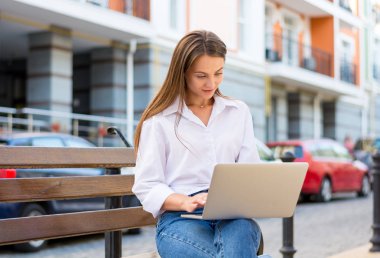 Genç güzel gülümseyen kadın bir dizüstü bilgisayar ile şehrin iş bölümünde bir bankta oturur. Genç güzel kadın, freelancer, yaz aylarında dizüstü bilgisayarda çalışır