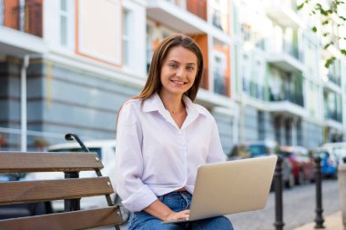 Genç güzel gülümseyen kadın bir dizüstü bilgisayar ile şehrin iş bölümünde bir bankta oturur. Genç güzel kadın, freelancer, yaz aylarında dizüstü bilgisayarda çalışır