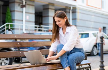 Genç güzel kadın ile bir laptop ve kulaklık kentin iş bölümünde bir bankta oturur. Genç güzel kadın, freelancer, yaz aylarında dizüstü bilgisayarda çalışır