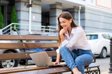 Genç güzel kadın ile bir laptop ve kulaklık kentin iş bölümünde bir bankta oturur. Genç güzel kadın, freelancer, yaz aylarında dizüstü bilgisayarda çalışır