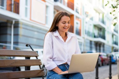 Genç ile bir dizüstü bilgisayar kadın gülüyor güzel şehrin iş bölümünde bir bankta oturur. Genç güzel kadın, freelancer, yaz aylarında dizüstü bilgisayarda çalışır