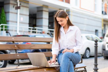 Genç güzel kadın ile bir laptop ve kulaklık kentin iş bölümünde bir bankta oturur. Genç güzel kadın, freelancer, yaz aylarında dizüstü bilgisayarda çalışır