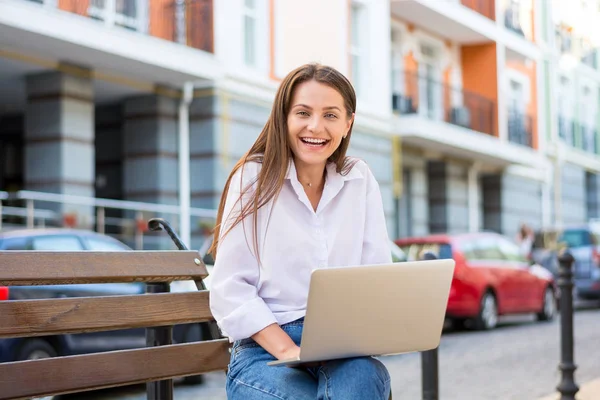 年轻美丽的笑女性与笔记本电脑坐在长椅上的商业部分城市 年轻美丽的女人 自由职业者 在笔记本电脑上夏天工作 — 图库照片