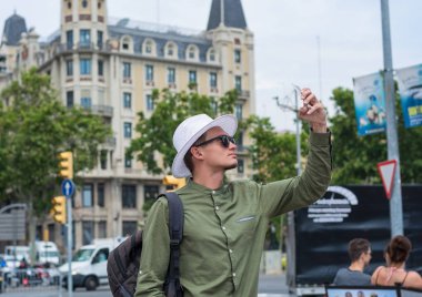 Genç bir adam, bir şapka ve bir sırt çantası ile ayakta turist ve smartphone merkezi Barselona, İspanya tarafından fotoğraf çeker