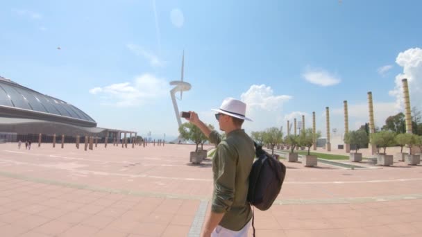 一个年轻男子 游客站在一个背包和帽子 并拍摄视频由智能手机附近的圣地亚哥卡拉特拉瓦和奥林匹克圆环 Anella Olmpica Montjuc Montjuc 的巴塞罗那 西班牙 — 图库视频影像